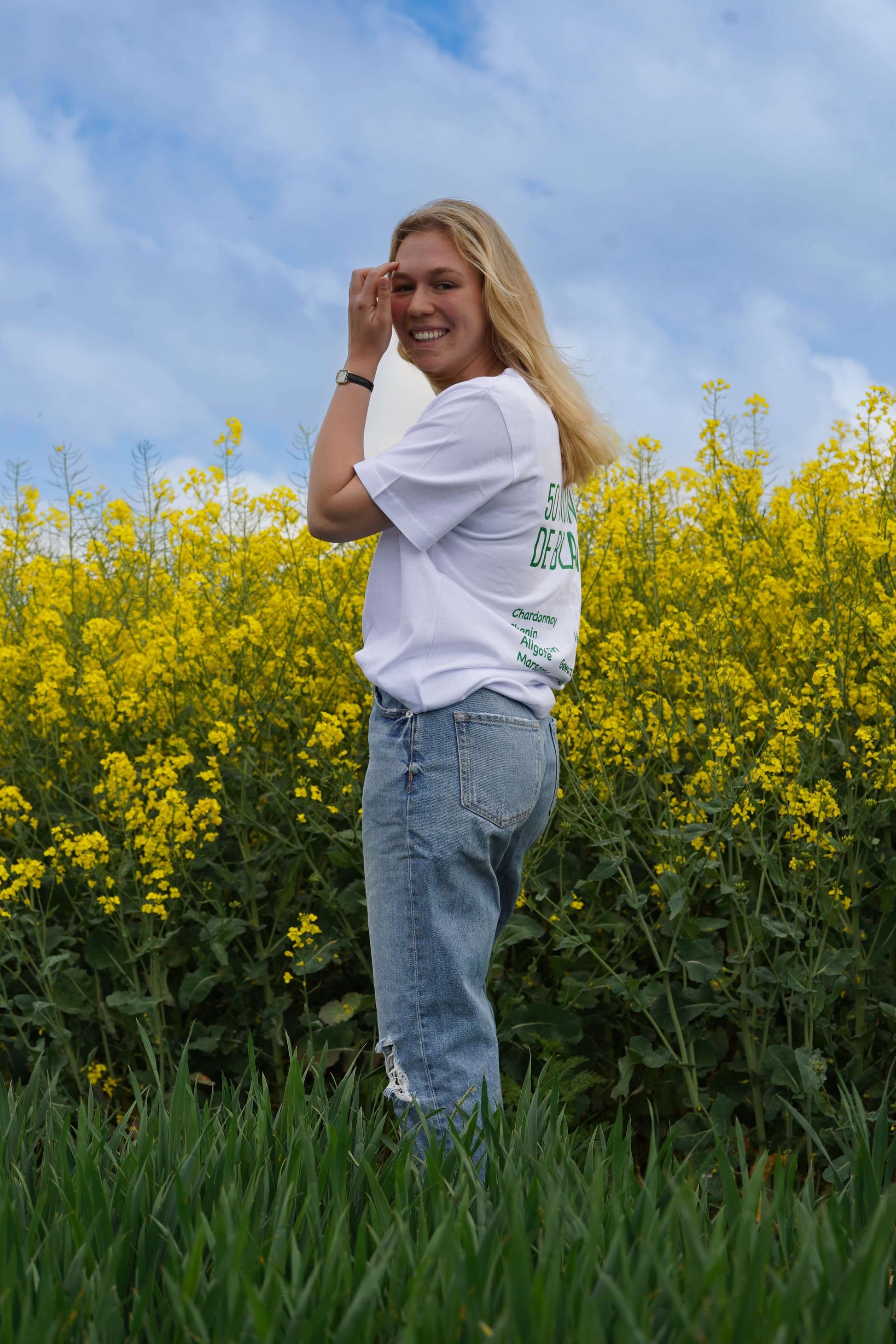 Femme souriante dans un t-shirt blanc "50 Nuances de Blancs" devant un champ de fleurs jaunes.