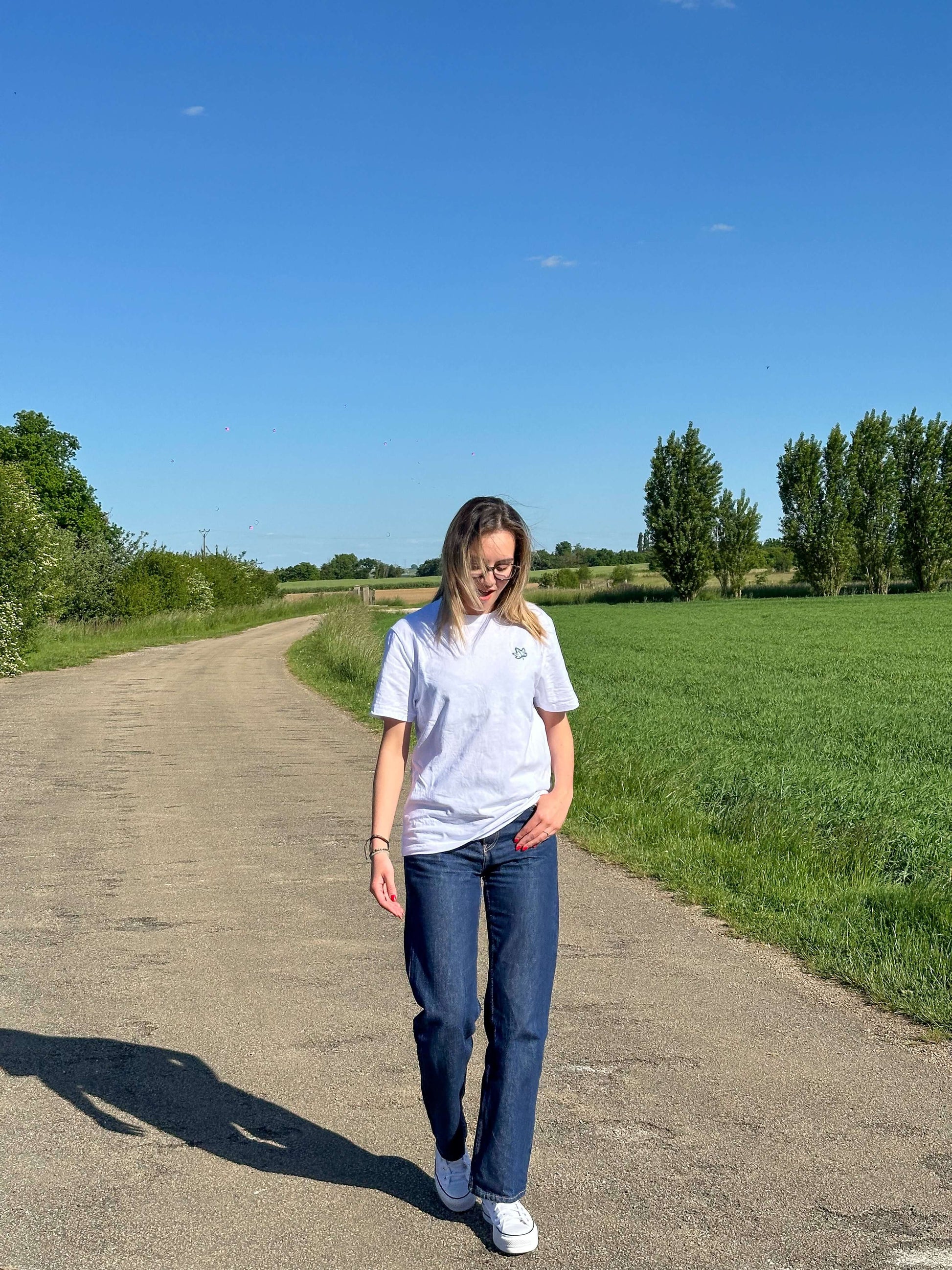 Woman walking on a country road wearing a casual grey T-shirt and jeans, surrounded by green fields and blue sky.