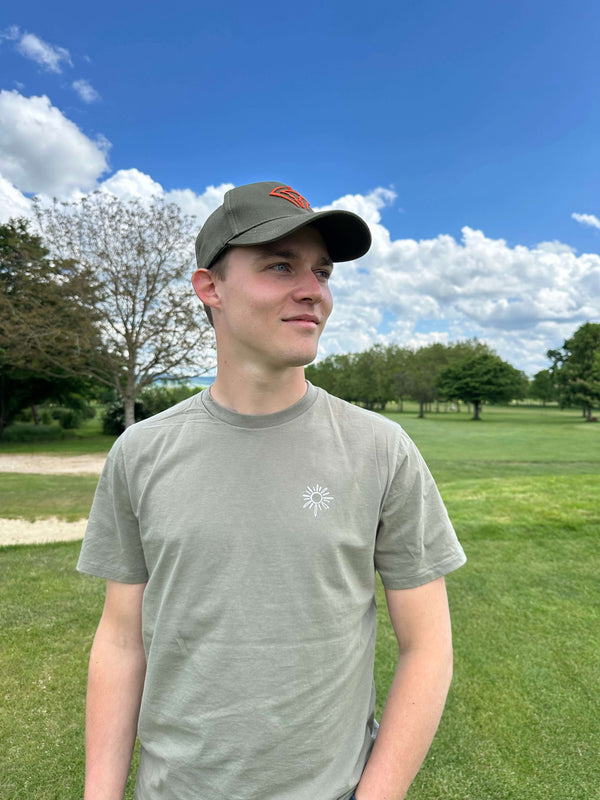 Young man wearing a casual olive cotton t-shirt and hat outdoors, smiling against a bright blue sky.