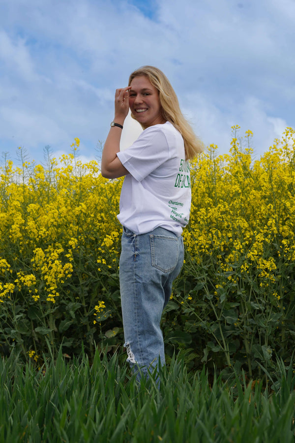 Femme souriante dans un t-shirt blanc "50 Nuances de Blancs" devant un champ de fleurs jaunes.