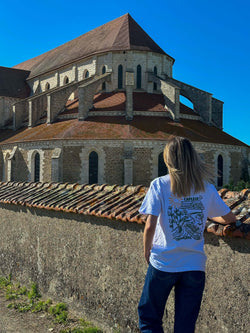 Image of Person wearing a white t-shirt with a design, standing by a historic building in a sunny landscape.