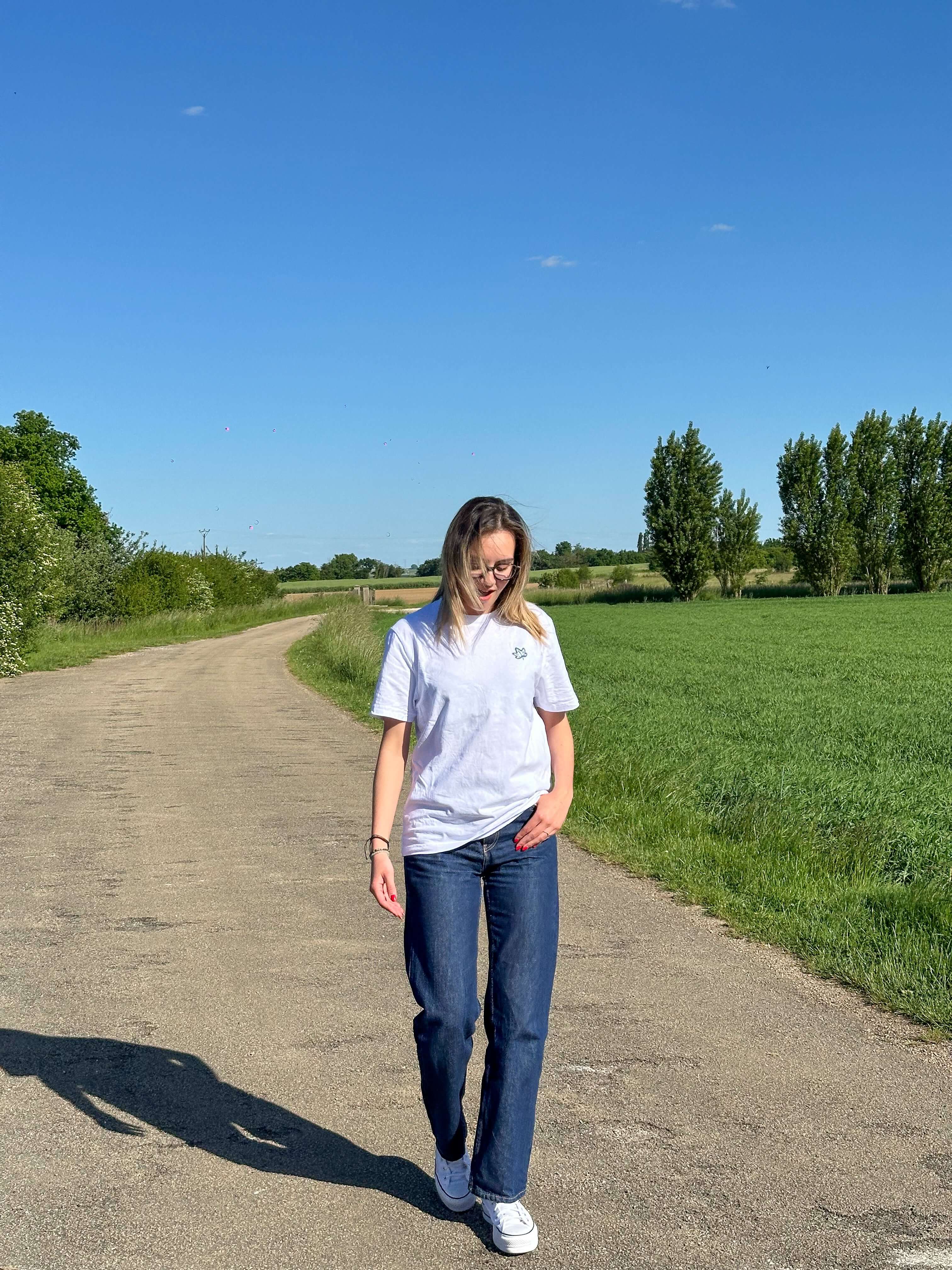 Image of Woman walking on a country road wearing a casual grey T-shirt and jeans, surrounded by green fields and blue sky.